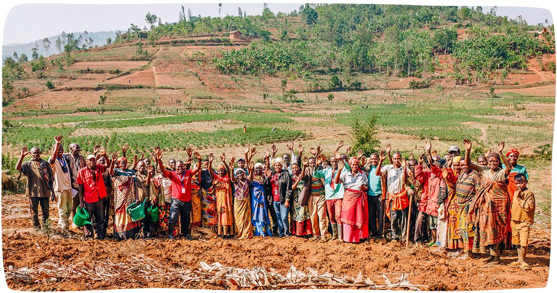 A large group of smiling Burundian people stand and wave in a field with a hillside in the background in Kabarore, Burundi